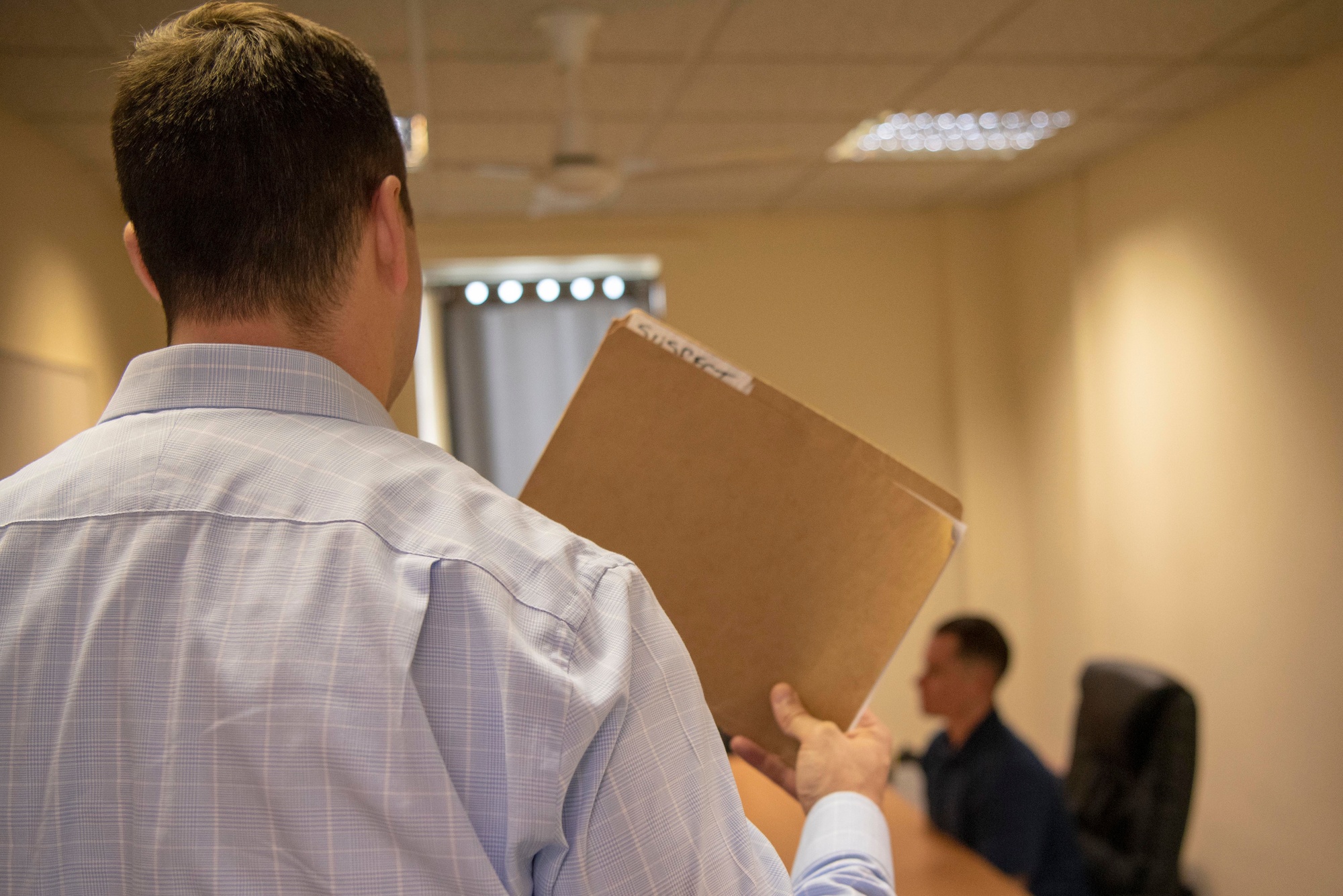 An investigator holds a case folder in the foreground of a simulated interview, with a seated participant out of focus.